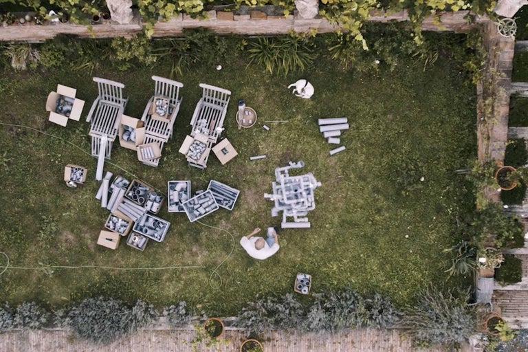 Letecký pohled na zahradu s osobou, zahradními židlemi, lepenkovými krabicemi, trubkami a psem