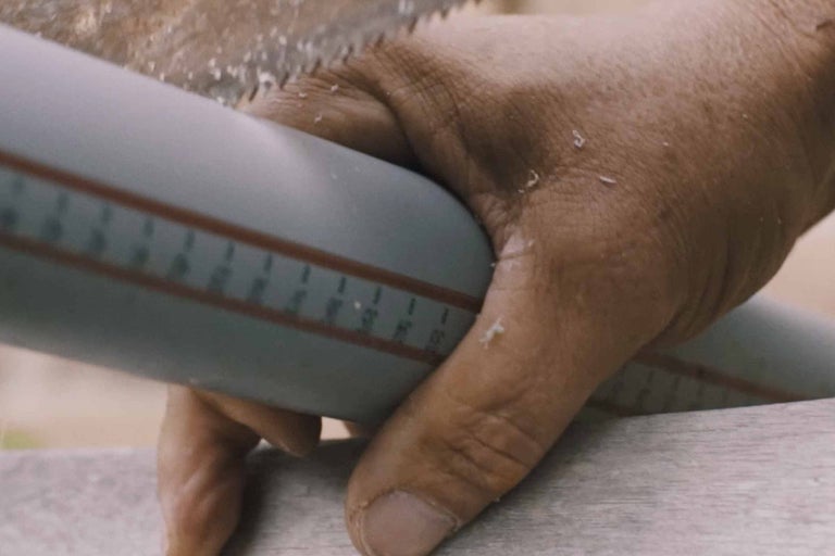 Detailní záběr muže řezajícího šedou plastovou trubku ruční pilou.