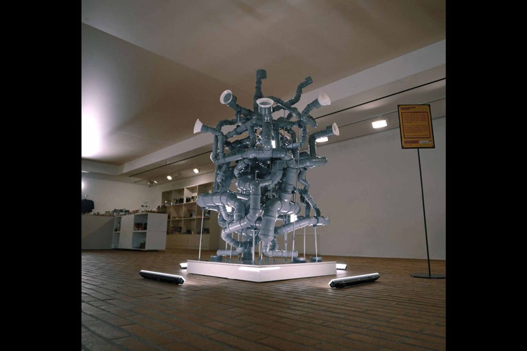 Socha z šedých plastových trubek ve výstavní místnosti.