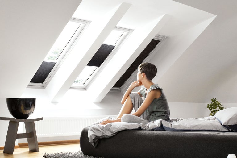 Světlá podkrovní místnost se střešními okny a zatemňovacími roletami, žena sedí na posteli
