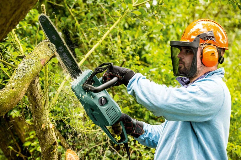 Osoba s ochrannými pomůckami používá elektrickou pilu k řezání větve.