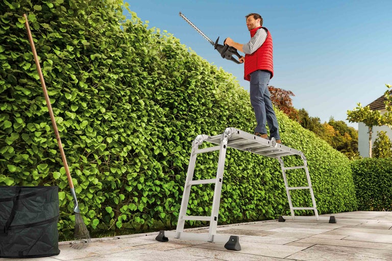Muž stojí na víceúčelovém žebříku a stříhá živý plot nůžkami na živý plot. Vedle stojí sběrač listí a hrábě.