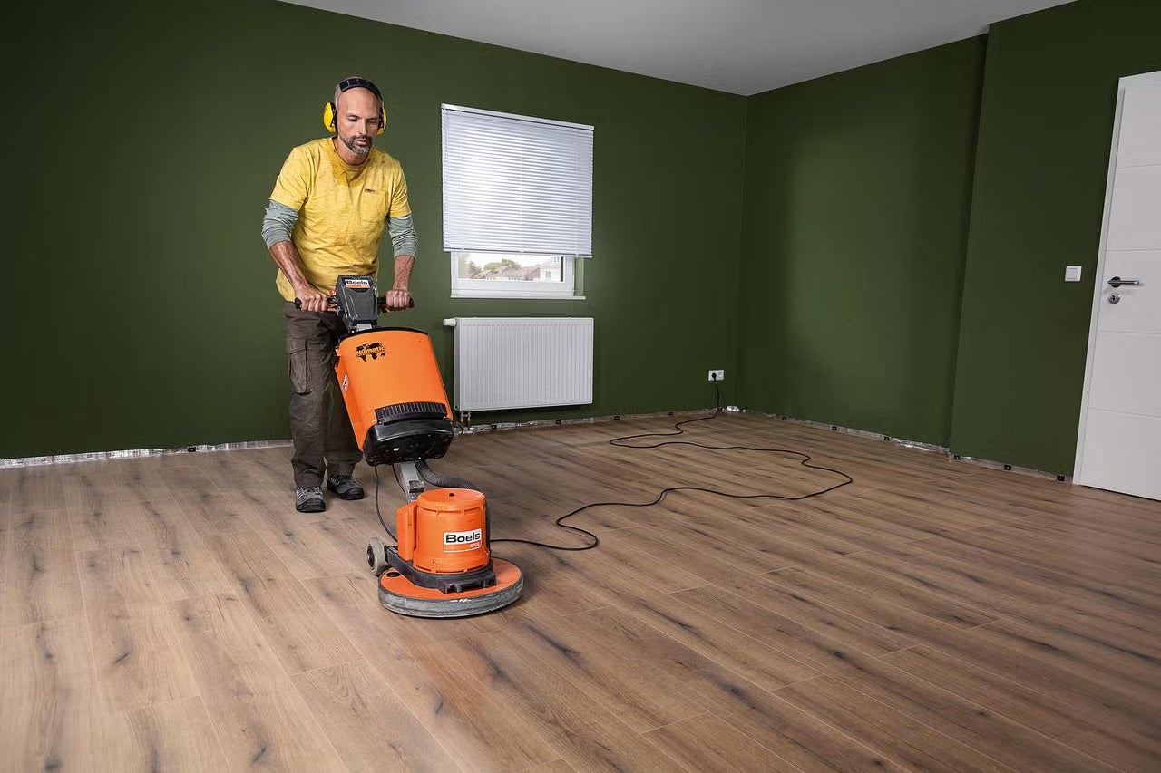 Muž s ochranou sluchu obsluhuje stroj na čištění podlah Boels na laminátové podlaze v jasné místnosti s oknem a radiátorem.