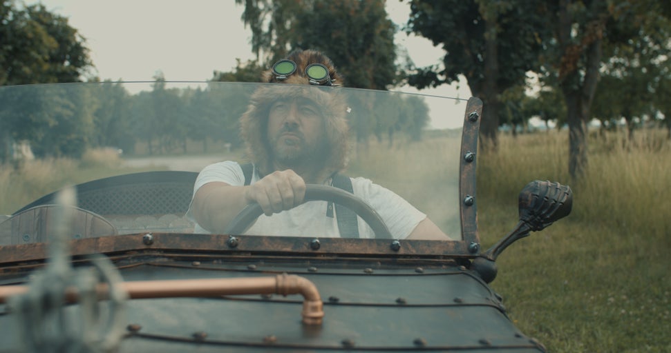 Muž s kožešinovou čepicí a brýlemi řídí auto v retro stylu.