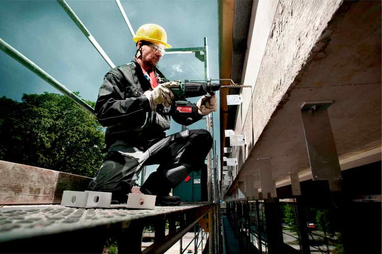 Stavební dělník na lešení vrtá s vrtacím kladivem do betonové stěny.