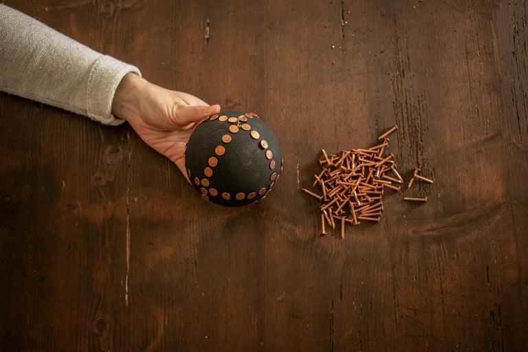 Ruka drží černou kouli s měděnými nýty vedle hromady hřebíků na dřevěném stole