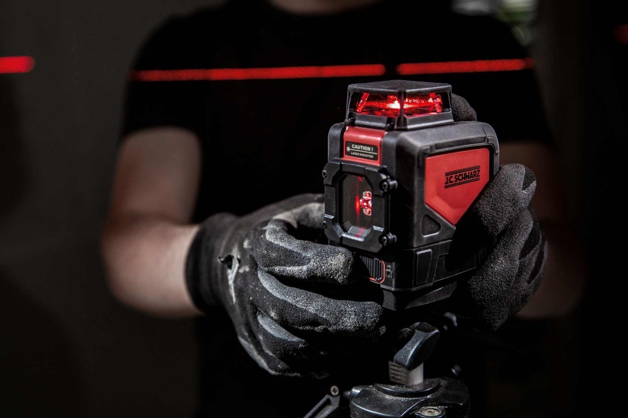 Osoba drží křížový liniový laser značky JC Schwarz, který promítá červené laserové čáry.
