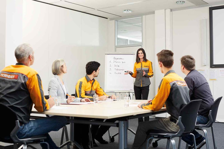 Zasedací místnost s flipchartem a skupinou lidí v pracovním oblečení během prezentace.