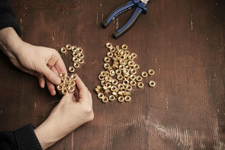 Osoba navléká mosazné matice drátem; na stole leží kleště.