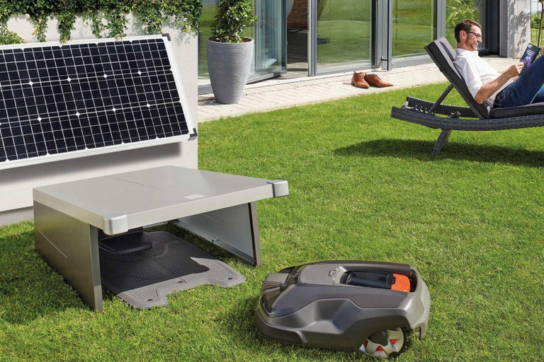 Zahradní scéna s robotickou sekačkou, nabíjecí stanicí, solárním panelem a mužem odpočívajícím na lehátku.