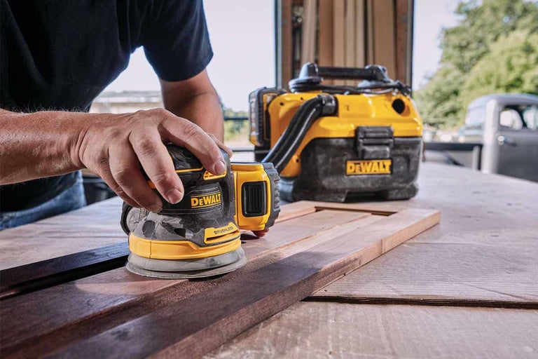 Osoba brousí dřevo excentrickou bruskou značky DeWalt. V pozadí je vysavač.