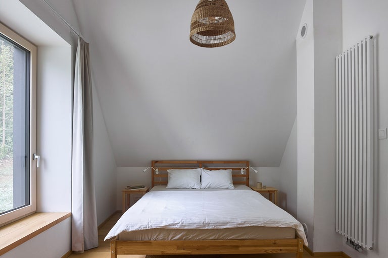 Ložnice s dřevěným rámem postele, nočními stolky, radiátorem, oknem a slámovou lampou