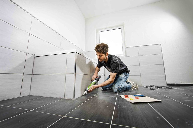 Řemeslník utěsňuje vanu kartuší v koupelně s šedými obklady.