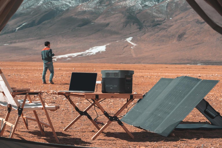 Venkovní scéna s notebookem, solárním panelem a napájecí stanicí EcoFlow na dřevěném stole v horské krajině.