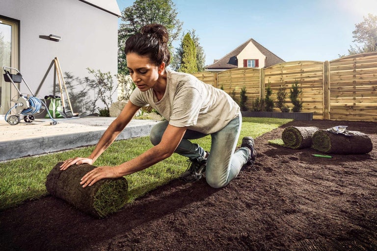 Žena pokládá travní koberec na zahradě před dřevěným plotem