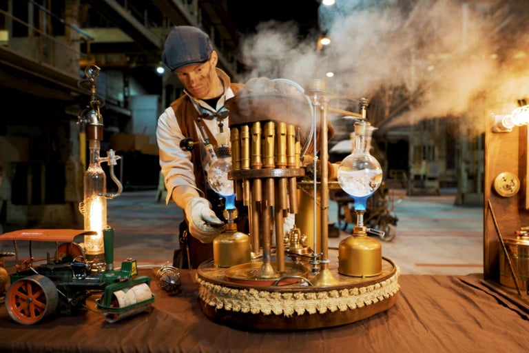 Muž ve steampunkovém oblečení obsluhuje parní stroj se zlatými prvky