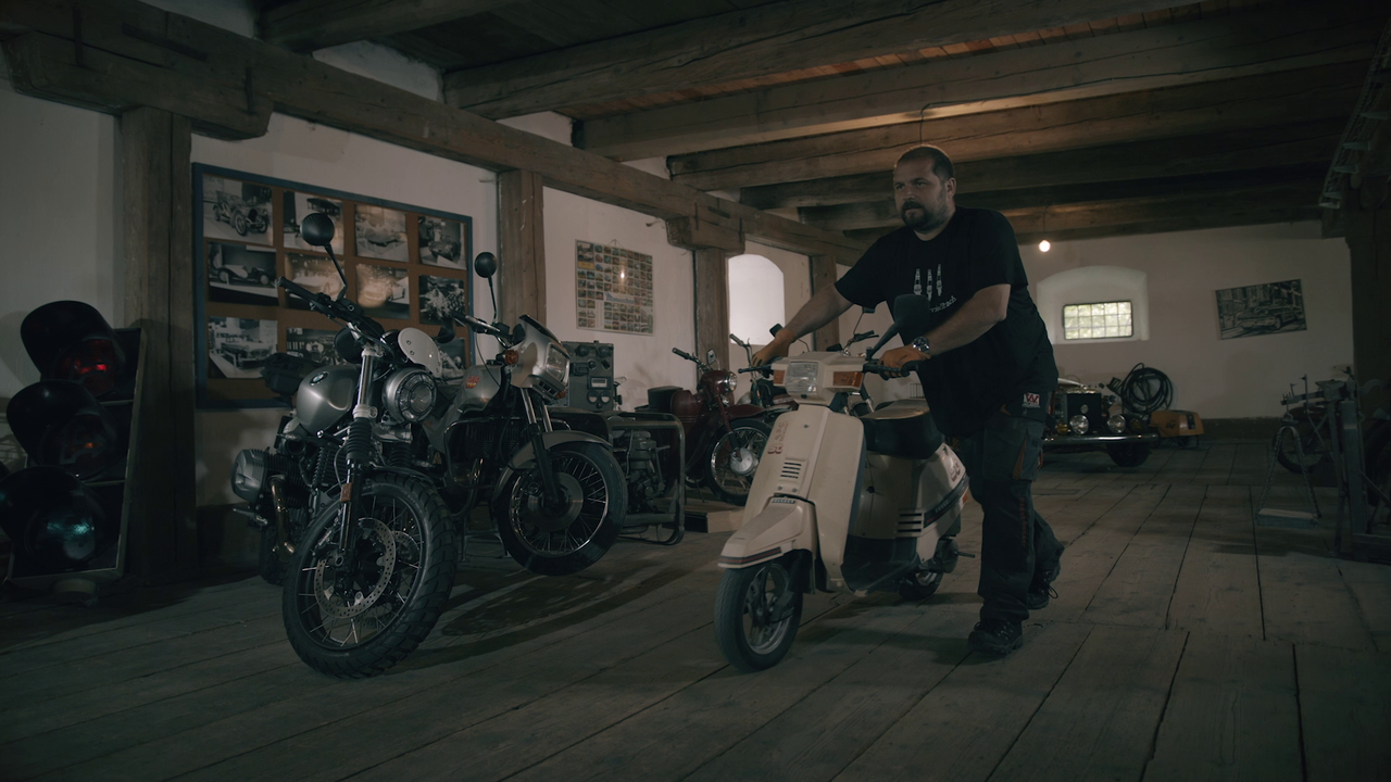 Muž tlačí skútr v dílně s různými motocykly
