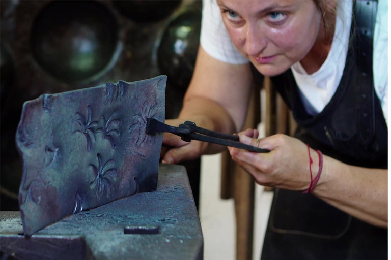 Kovárna zpracovává kus kovu s květinovým vzorem pomocí kleští na kovadlině.