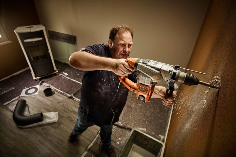 Muž vrtá vrtacím kladivem do zdi v místnosti. V pozadí jsou vidět kamna a kamnové roury.