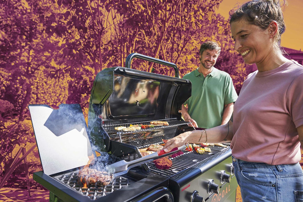 Žena griluje maso a zeleninu na velkém plynovém grilu na světlé zahradě, zatímco muž v pozadí přihlíží.