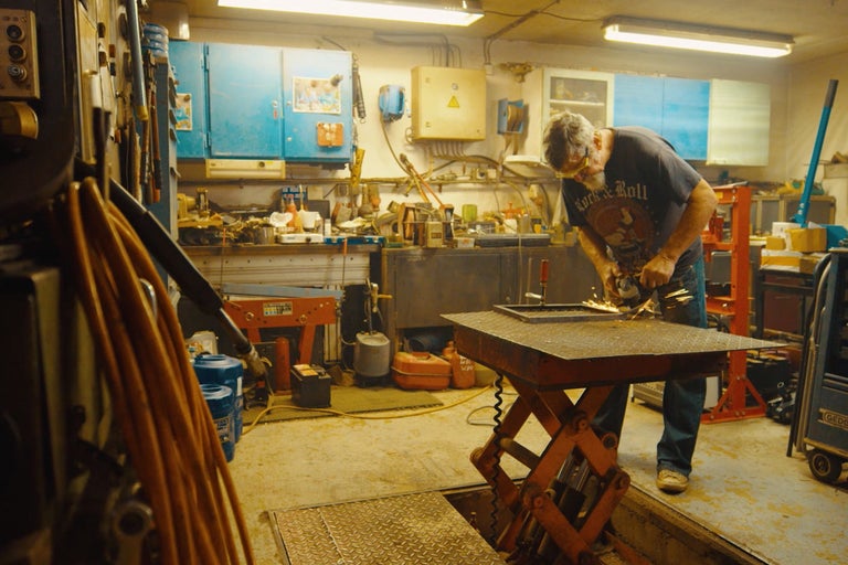 Dílna s mužem, který brousí kov, pracovní stůl a nářadí.