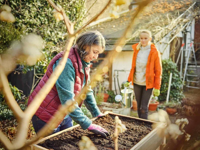 Dvě ženy pracují na zahradě s vyvýšeným záhonem, konví a žebříkem.