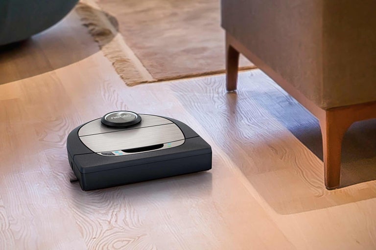 Logo Neato. Robotický vysavač na dřevěné podlaze vedle koberce a židle.