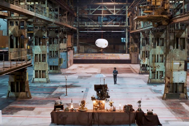 Interiér tovární haly s mužem pod meteorologickým balónem v dálce a stolem s dekoracemi v popředí.