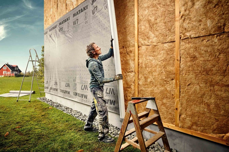 Muž montuje fasádní fólii na stěnu domu s izolační vatou.