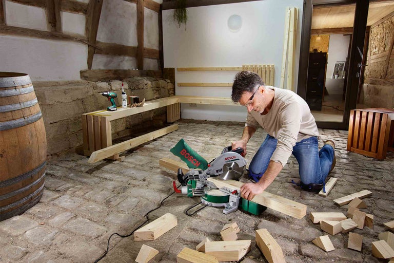 Muž řeže dřevo pokosovou pilou v dílně. V pozadí je vidět sud, dřevo a nářadí.