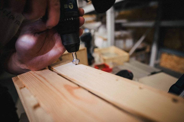 Ruka drží vrtačku, která opracovává kus dřeva.