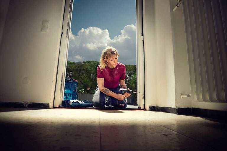 Žena klečí před otevřenými dveřmi a pracuje s akumulátorovým šroubovákem. V pozadí je vidět kufr s nářadím.