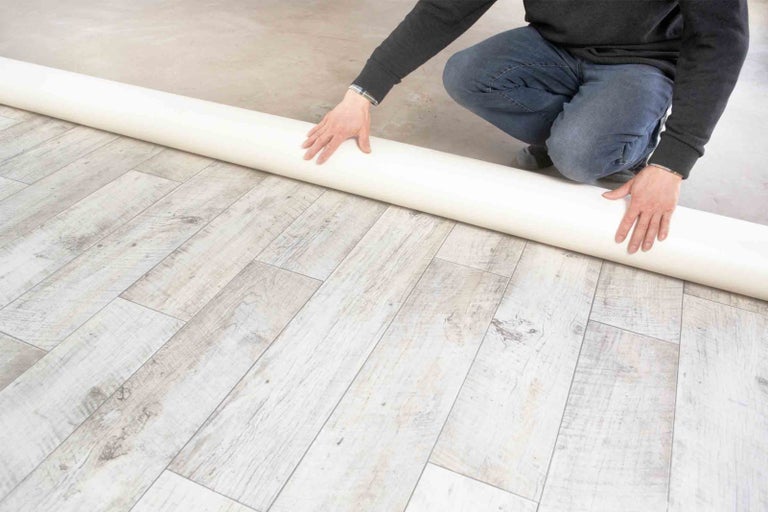 Osoba pokládá roli bílé podlahové krytiny na dřevěnou podlahu