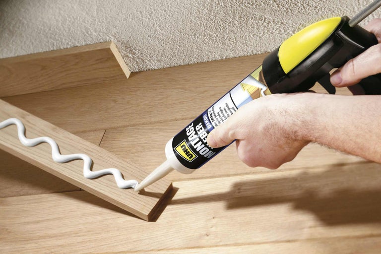Řemeslník nanáší montážní lepidlo na dřevěnou lištu pomocí kartuše.