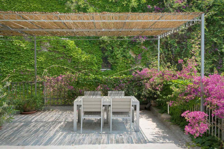 Terasa s jídelním stolem a židlemi pod pergolou, obklopená bujnou zelení.