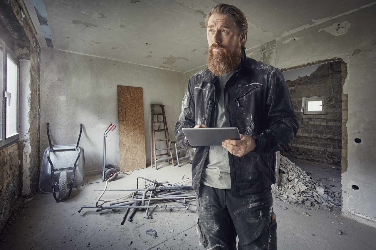 Řemeslník s tabletem v místnosti s hrubou stavbou, nářadím a stavebními materiály.