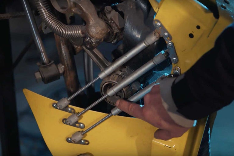 Detailní záběr žlutého stroje s mechanickými částmi a prstem ukazujícím na součástku