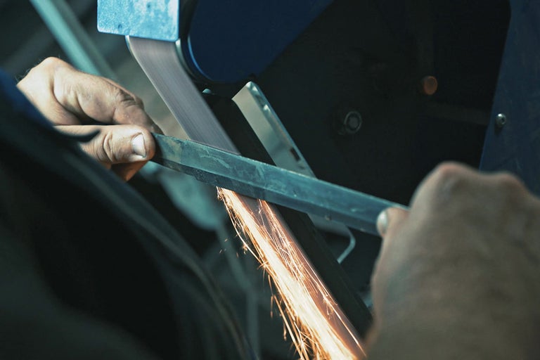 Muž brousí kov pásovou bruskou, létají jiskry.