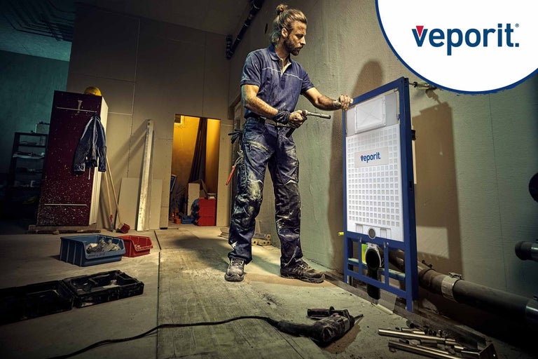 Řemeslník montuje splachovací nádržku před stěnu v nedokončené místnosti. Logo Veporit je viditelné.