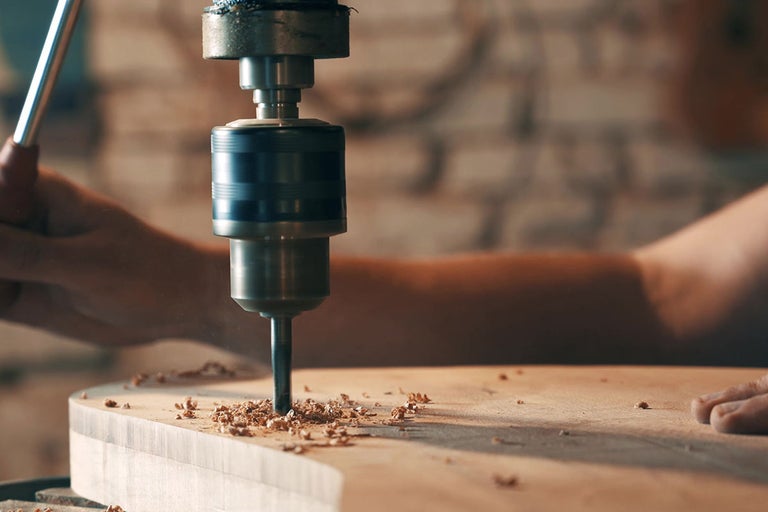 Detail stolní vrtačky vrtající do kusu dřeva.