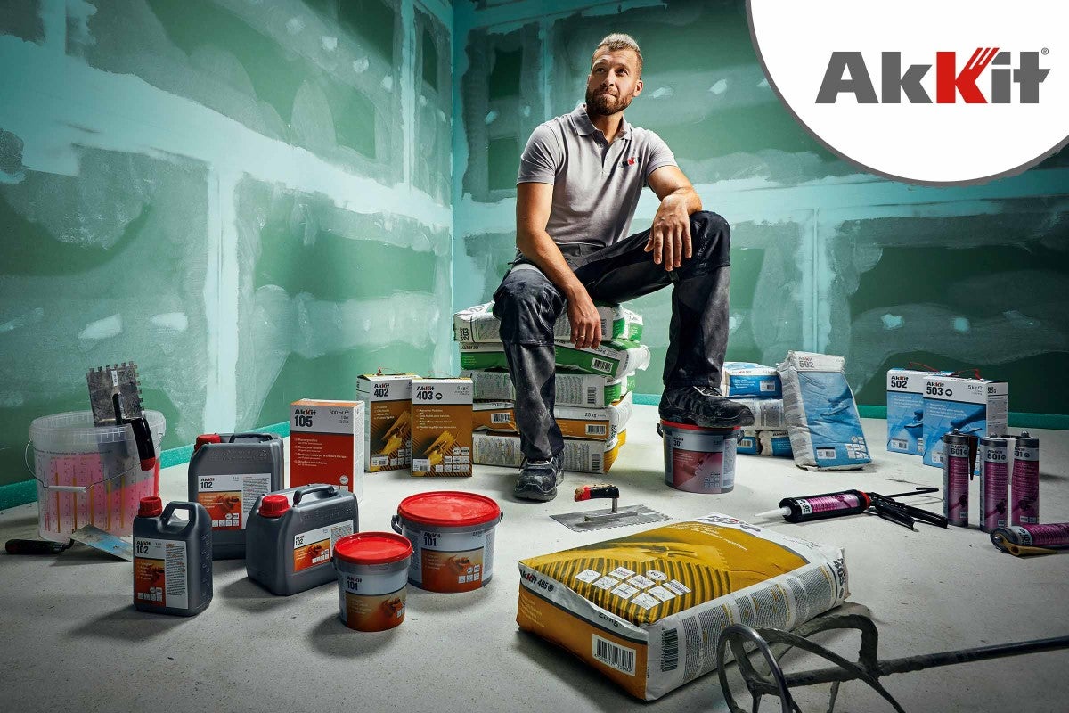Muž sedí na pytlích s produkty Akkit v místnosti s omítnutými stěnami, obklopený různými produkty Akkit, jako jsou lepidla a tmely
