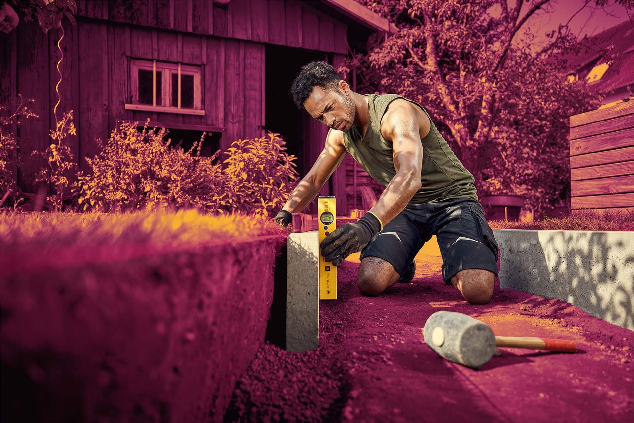 Muž na zahradě vyrovnává betonový obrubník pomocí vodováhy, v blízkosti leží gumová palice.