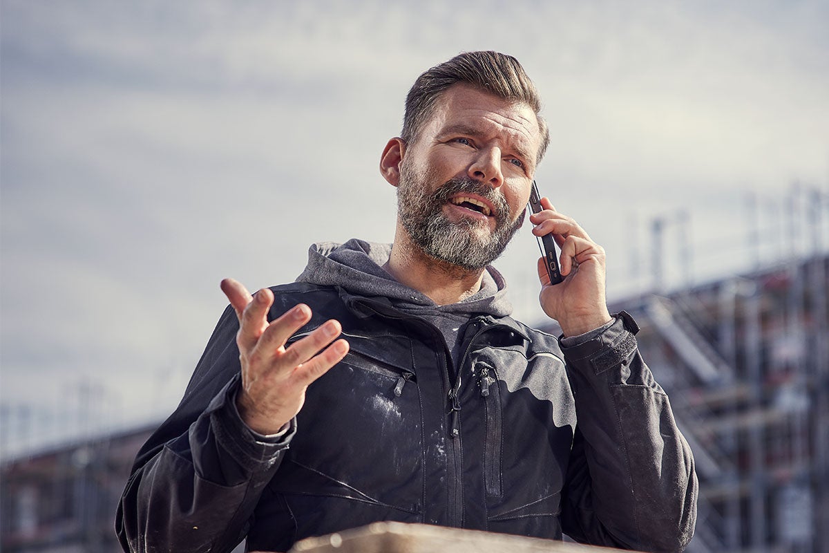 Vousatý muž v tmavé pracovní bundě a mikině telefonuje na staveništi s chytrým telefonem a přitom gestikuluje.