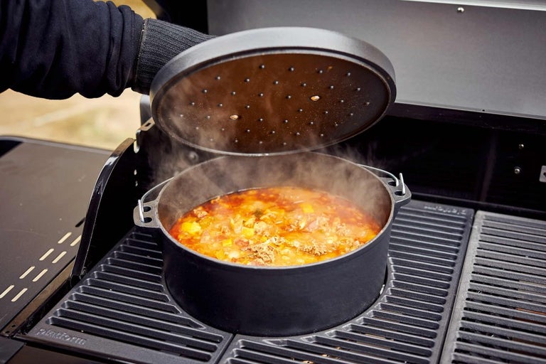 Holandská trouba s víkem stojí na grilu a je plná jídla.