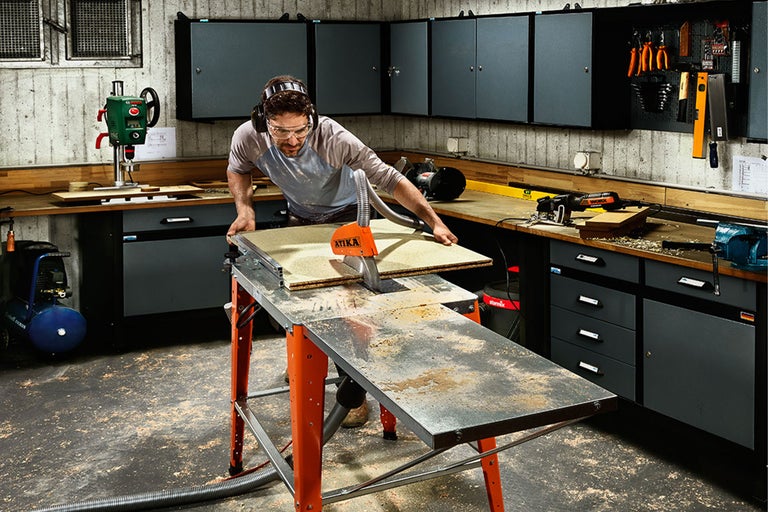 Dílenská scéna se stolní kotoučovou pilou, pracovní stůl a osoba s ochranou sluchu a ochrannými brýlemi, která řeže kus dřeva.