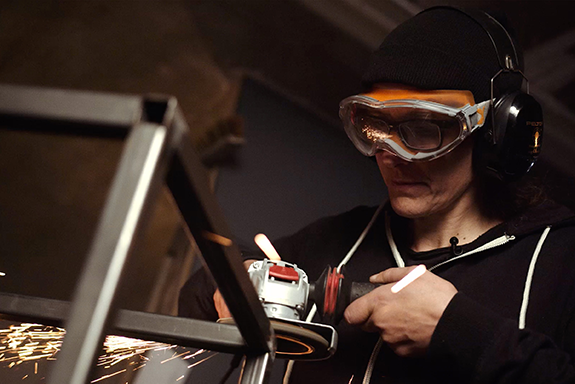 Osoba brousí kov pomocí brusky, přičemž má ochranné brýle a chrániče sluchu.