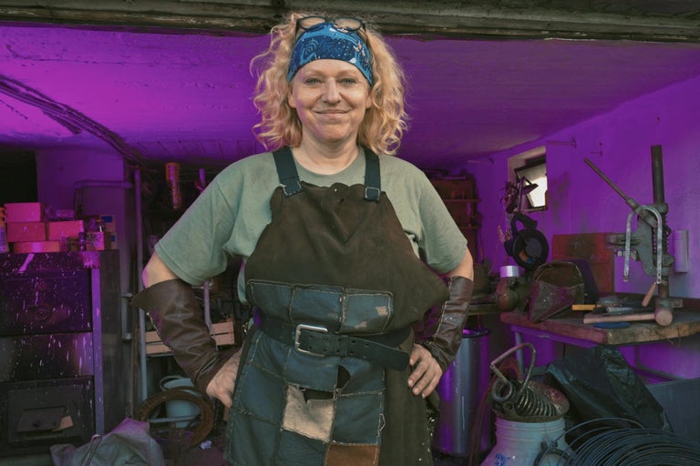 Žena nosí zástěru, kožené rukavice a čelenku v dílně