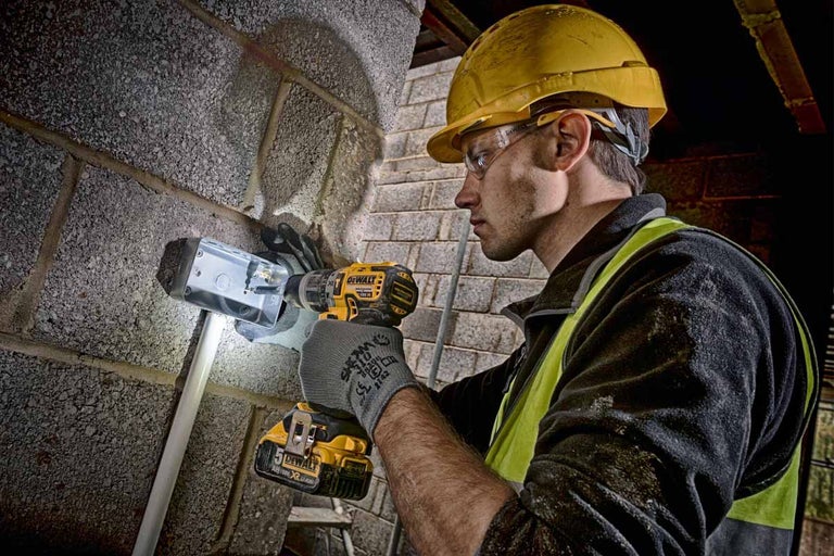 Řemeslník se žlutou helmou a ochrannými brýlemi vrtá akumulátorovým vrtacím šroubovákem otvor do zdi pro odbočnou krabici.