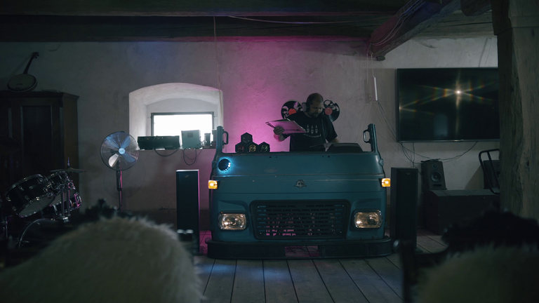 Interiér s DJ pultem vyrobeným z přední části modrého nákladního automobilu, doplněný DJ vybavením, reproduktory a mužem pouštějícím desky.