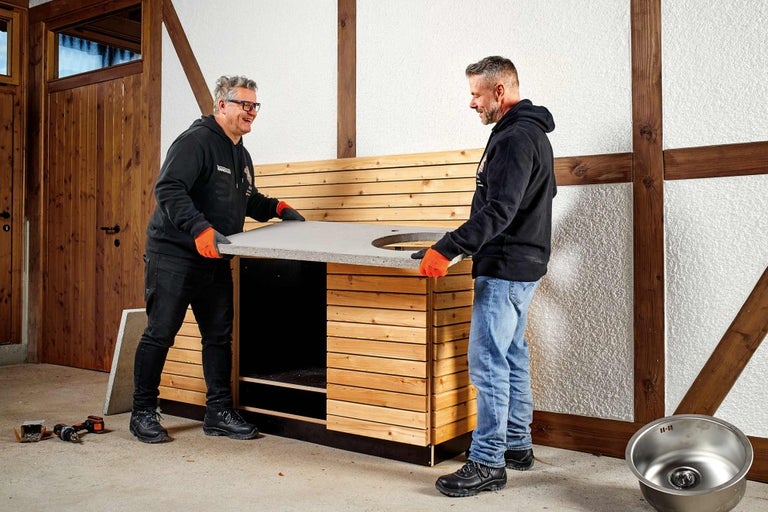 Dvě osoby montují pracovní desku s výřezem pro dřez na dřevěnou venkovní kuchyni.